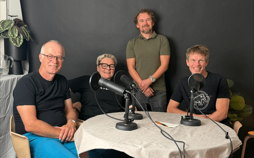 Bilde av programlederne og gjestene i studio. De sitter rundt et bord med podcastmikrofonene rundt seg
